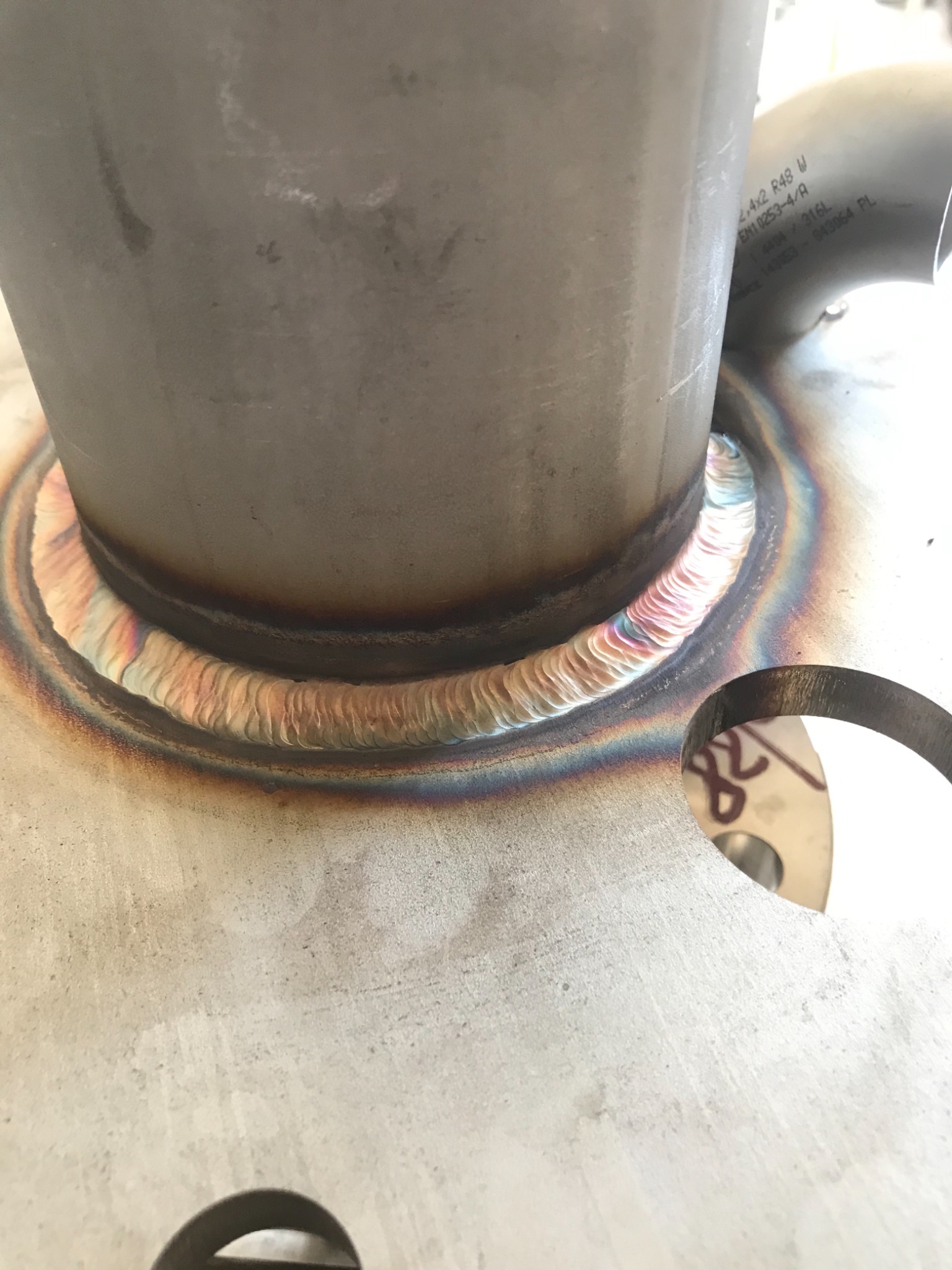 Szczegółowe ujęcie spoiny TIG łączącej rurę ze stalową płytą, widoczne tęczowe przebarwienia termiczne na spoinie.