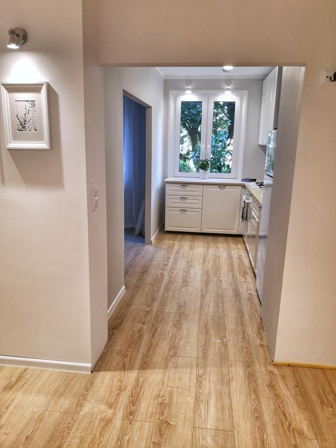 Jasne panele podłogowe imitujące drewno w korytarzu prowadzącym do kuchni z białymi meblami i oknem z widokiem na zieleń.