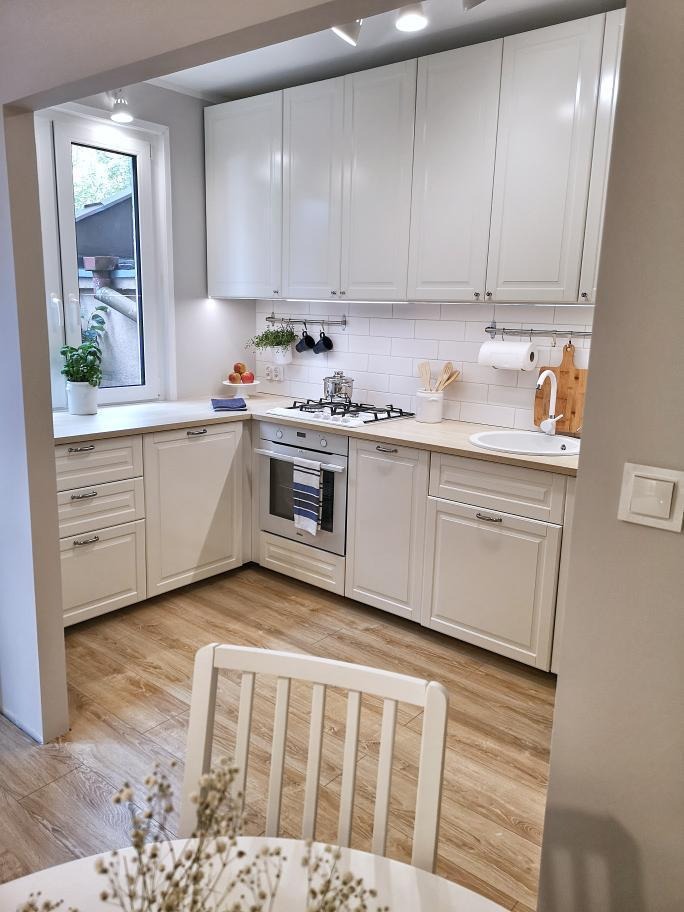 Jasna kuchnia w stylu skandynawskim z drewnianą podłogą i białymi szafkami, widok na okno z rośliną i fragment białego krzesła na pierwszym planie.