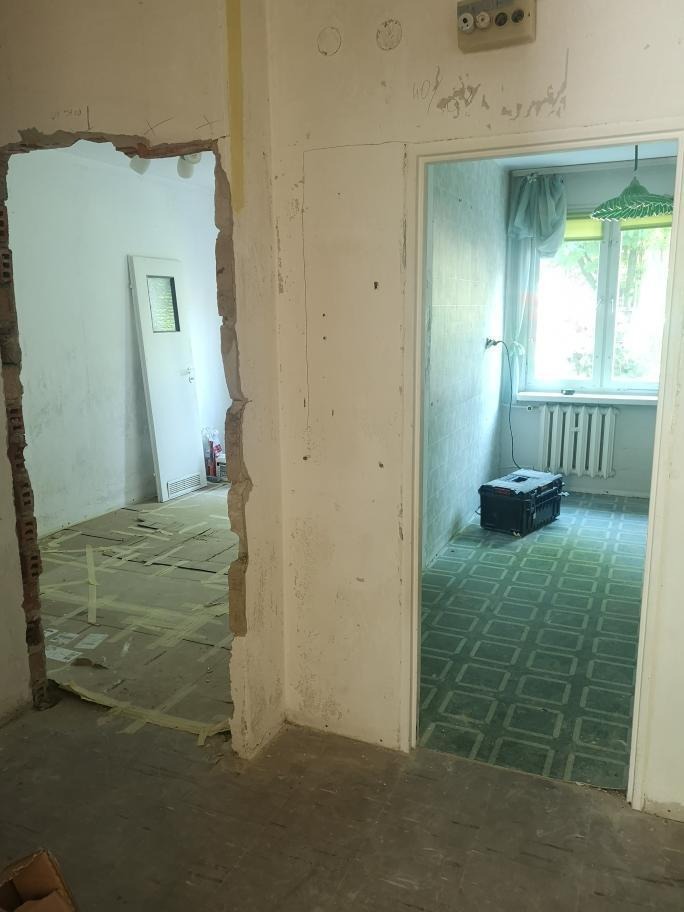 Widok na dwa pomieszczenia w trakcie prac: w jednym zerwane podłogi i gołe ściany, w drugim stara glazura i skrzynia narzędziowa.