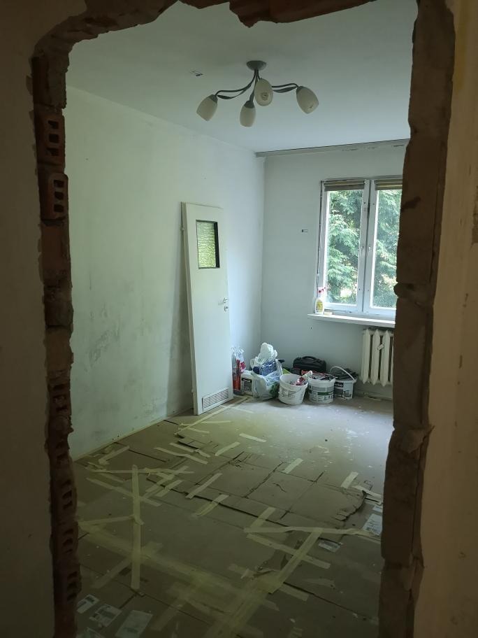 Pokój w trakcie prac przygotowawczych do remontu, podłoga zabezpieczona kartonem i taśmą, widoczne wiadra z farbą i narzędzia przy oknie, surowy otwór drzwiowy z fragmentami cegieł.