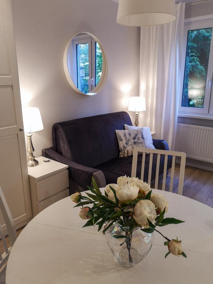 Jasne wnętrze pokoju dziennego z szarą sofą, okrągłym lustrem i białym okrągłym stołem z bukietem białych piwonii w szklanym wazonie.