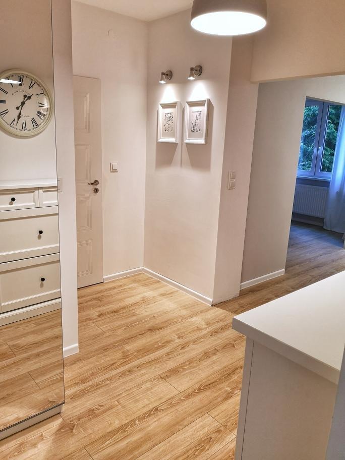 Jasne panele podłogowe w korytarzu, białe ściany z dekoracjami, widoczny fragment szafy z lustrem i zegarem.