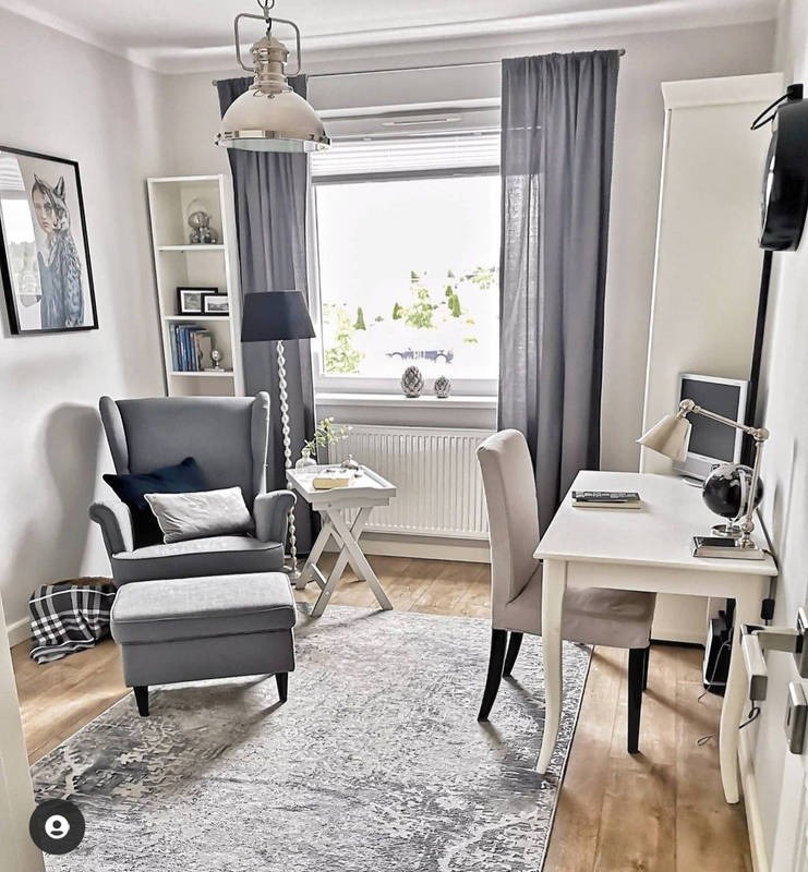 Jasne wnętrze pokoju z szarym fotelem, podnóżkiem i dywanem w stylu vintage, białe biurko z lampką, okno z szarymi zasłonami, lampa wisząca w stylu industrialnym, drewniana podłoga.