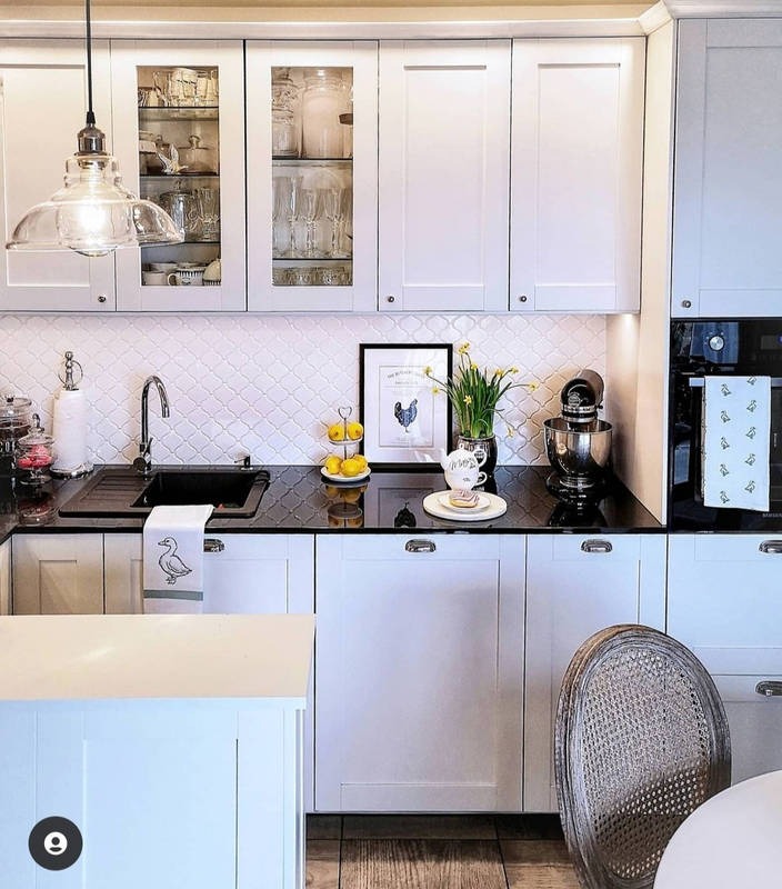 Elegancka, jasna kuchnia z czarnym blatem, białymi szafkami z przeszkleniami, mozaikową ścianą i dekoracyjnymi dodatkami, takimi jak obrazek z kogutem i cytryny na paterze.
