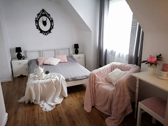 Jasna sypialnia na poddaszu z łóżkiem, stolikami nocnymi, fotelem obitym różowym pluszem i dekoracyjnym czarnym ornamentem na ścianie.