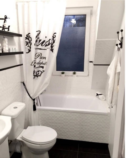 Biała łazienka z czarnymi akcentami: czarne półki z kosmetykami, zasłona prysznicowa z napisem 'Paris', czarne płytki podłogowe i biała wanna z dekoracyjnymi panelami. Na parapecie okna dwie małe...