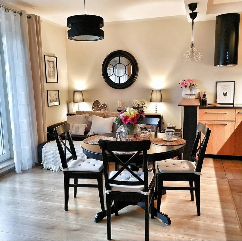 Jasne wnętrze mieszkania z okrągłym stołem jadalnym z czarnymi krzesłami, kwiatami w wazonie i widokiem na salon z sofą i dekoracjami, oświetlone lampami i wiszącymi lampami.