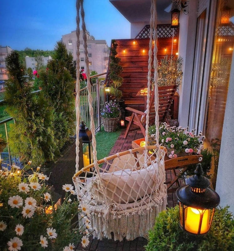 Przytulnie urządzony balkon z drewnianą podłogą, huśtawką hamakową z makramy, lampionami i roślinami w donicach, oświetlony girlandami świetlnymi, z widokiem na budynki mieszkalne w tle.