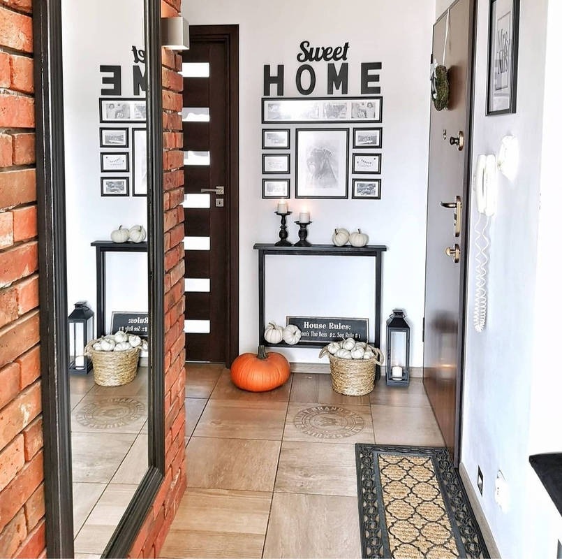Aranżacja przedpokoju z ceglaną ścianą, dużym lustrem, drewnianymi drzwiami z przeszkleniami, ceramiczną podłogą, dekoracjami w postaci dywanika, lampionów, koszy z małymi dyniami i napisem 'Sweet...