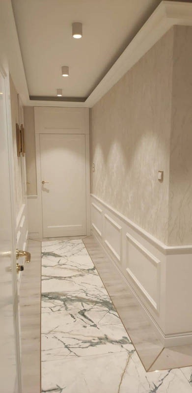 Wnętrze korytarza wykończonego w jasnych barwach z marmurową posadzką i listwami dekoracyjnymi na ścianach, oświetlonego punktowymi lampami sufitowymi.