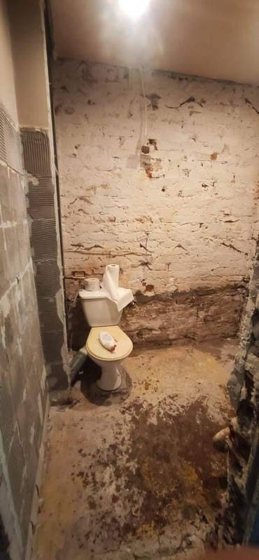 Łazienka w trakcie demontażu: stara toaleta z papierem toaletowym i środkiem czyszczącym na desce, ściany z odsłoniętą cegłą i resztkami płytek, zabrudzona podłoga.