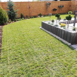 Dany Garden - Zadbany trawnik z rabatami kwiatowymi i ozdobnymi krzewami, obrzeża wykończone korą, podwyższona rabata z szarej kostki wypełniona białym grysem i miniaturowymi drzewkami, drewniany płot w tle.