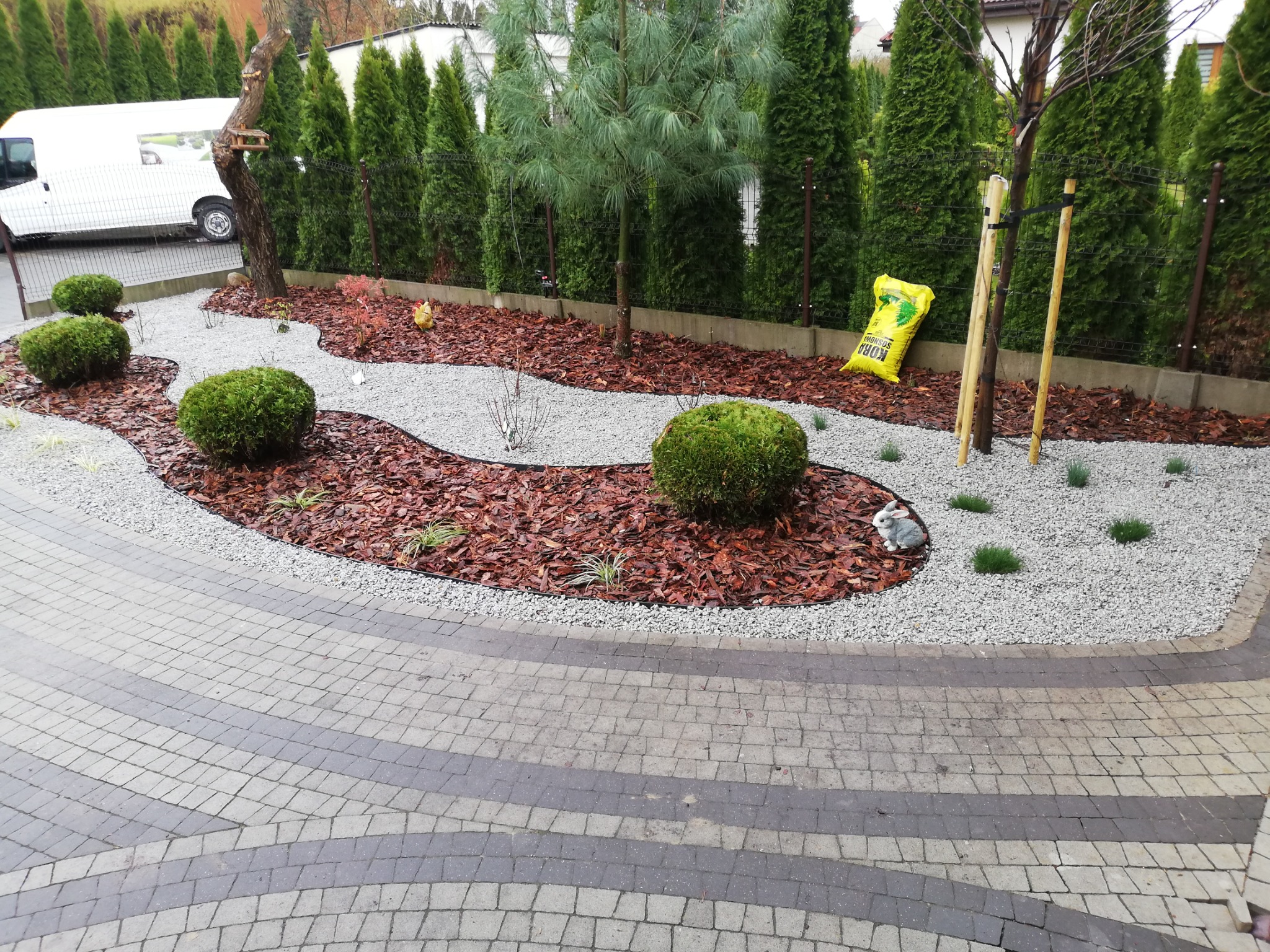 Świeżo zaaranżowany ogród z krzewami, korą, drobnym żwirem i figurką królika, widoczne tuje w tle i częściowo kostka brukowa na pierwszym planie.