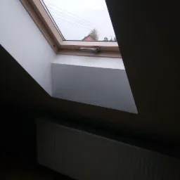 Poddasze z oknem dachowym z widokiem na zewnątrz, białe wykończenie wnęki okiennej, grzejnik na podłodze z paneli.