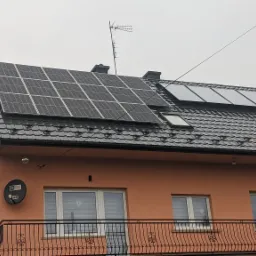 Instalacja paneli fotowoltaicznych na dachu domu jednorodzinnego w Krakowie, widoczne panele w dwóch sekcjach, dach pokryty blachodachówką, antena telewizyjna na szczycie, pochmurne niebo.