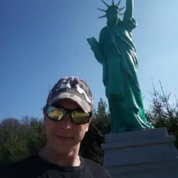 Mężczyzna w okularach przeciwsłonecznych i czapce moro robiący selfie na tle zielonej repliki Statuy Wolności na betonowym cokole, z widocznymi drzewami w tle i błękitnym niebem.