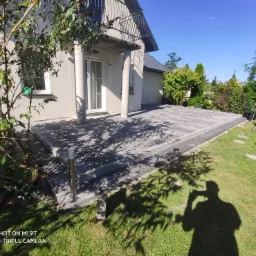 Nowoczesny taras z szarej kostki brukowej ułożonej na podwyższeniu, widok z trawnika, cień drzewa i sylwetka fotografa.