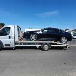 Srebrna laweta z czarnym samochodem osobowym na platformie, w tle budynek stacji benzynowej i ceglany budynek.