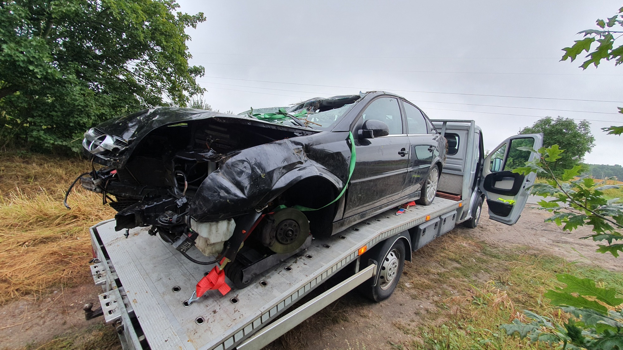 Czarny samochód osobowy po wypadku, z poważnymi uszkodzeniami przodu, zabezpieczony zielonym pasem, załadowany na lawetę w plenerze, widoczne otwarte drzwi lawety.