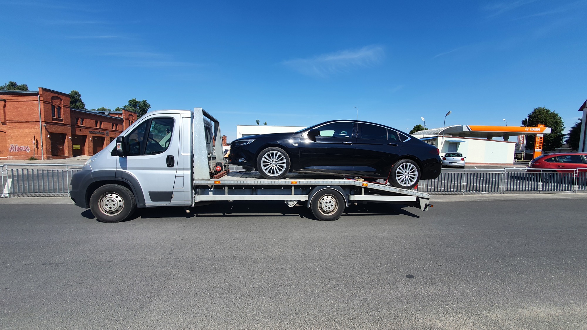 Srebrna laweta z czarnym samochodem osobowym na platformie, w tle budynek stacji benzynowej i ceglany budynek.