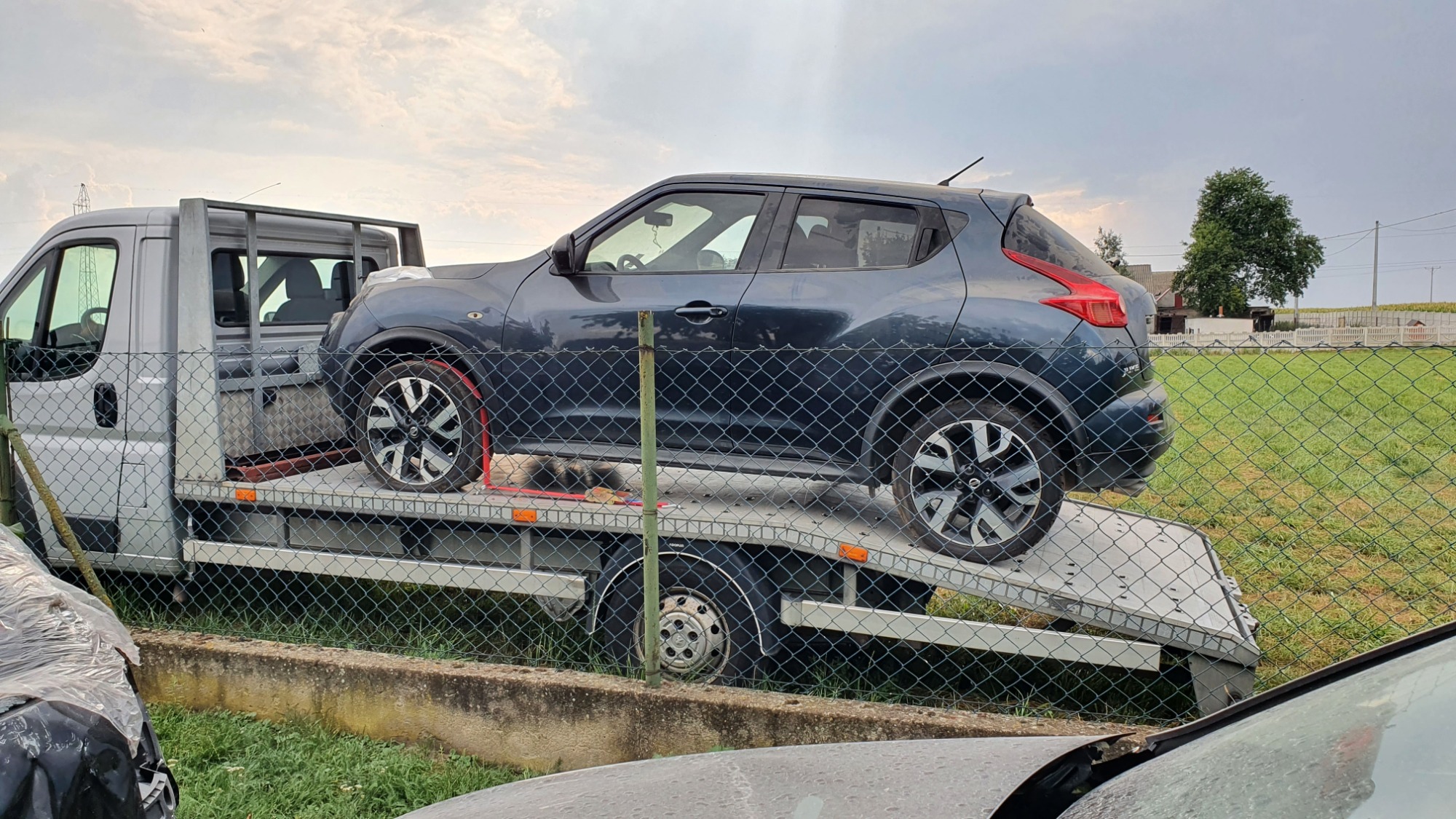 Szary Nissan Juke zabezpieczony na platformie lawety, widoczny przez siatkę ogrodzeniową, z fragmentem srebrnego busa holującego w tle.
