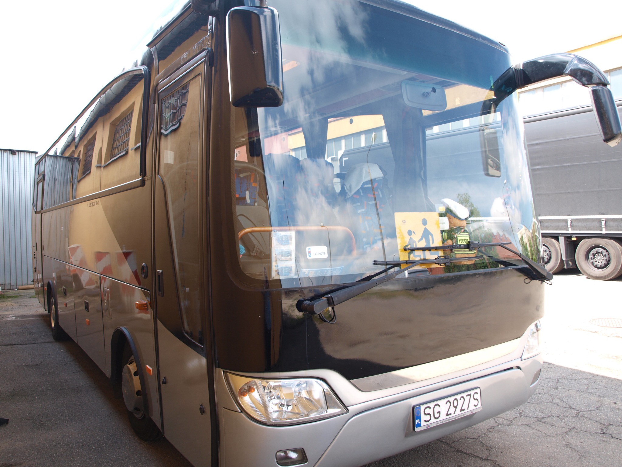 Przód szaro-czarnego autokaru turystycznego z widoczną tablicą rejestracyjną SG 2927S i odbiciami w przedniej szybie, stojącego na zewnątrz budynku.