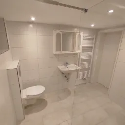 Jasna łazienka z beżowymi płytkami podłogowymi i białymi płytkami ściennymi, wisząca toaleta z zabudowaną spłuczką, umywalka z szafką lustrzaną, grzejnik drabinkowy i szklana ścianka prysznicowa.