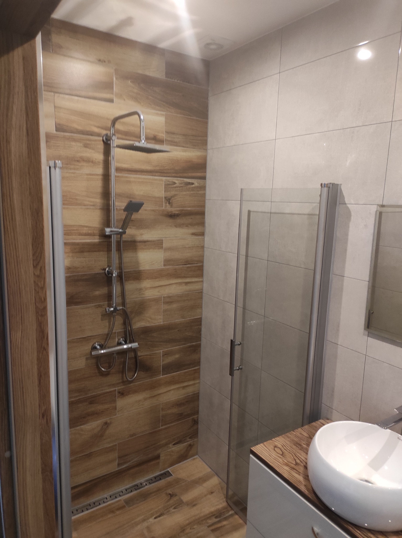 Nowoczesna kabina prysznicowa z deszczownicą i panelami imitującymi drewno, umywalka nablatowa na szafce z blatem w odcieniu drewna, jasne płytki na ścianach.