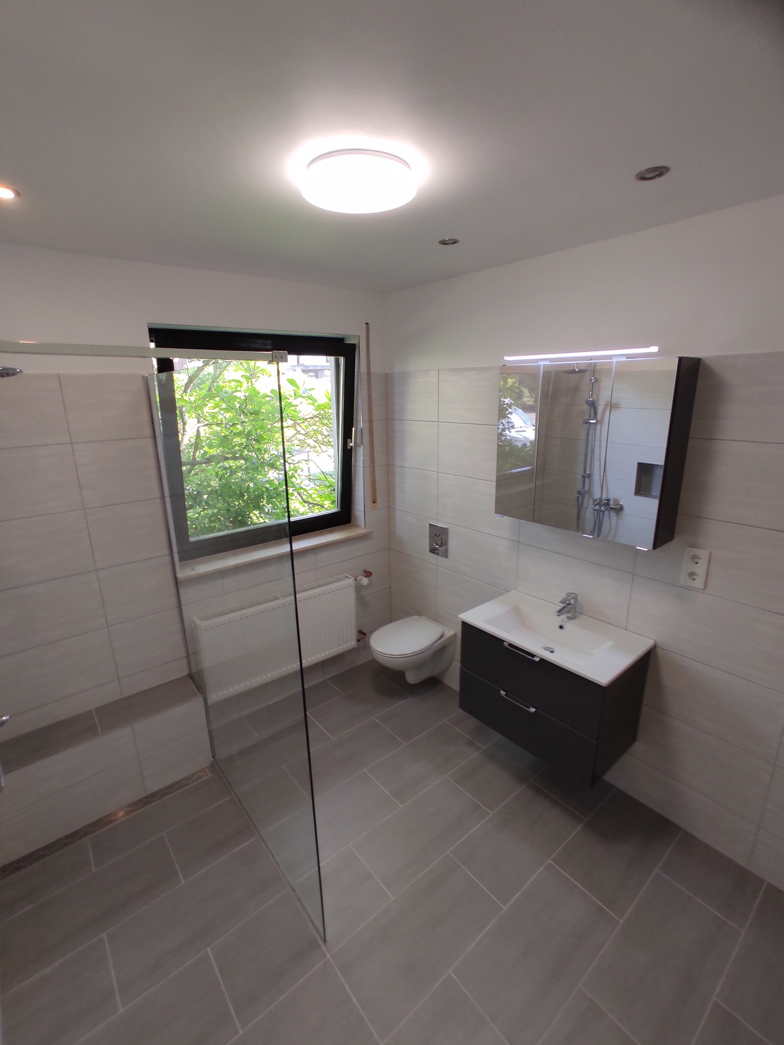 Nowoczesna łazienka z szarymi płytkami, kabiną prysznicową typu walk-in i umywalką z szafką w kolorze antracytowym, widok z góry, okno z widokiem na zieleń.