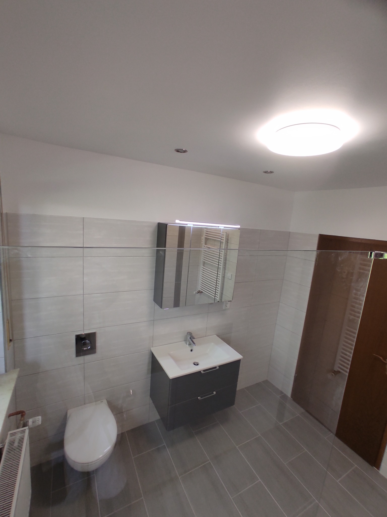 Nowoczesna łazienka z szarymi płytkami, podwieszaną toaletą, szafką z umywalką i lustrem z oświetleniem LED, widoczny grzejnik drabinkowy oraz drzwi z hydropowłoką.