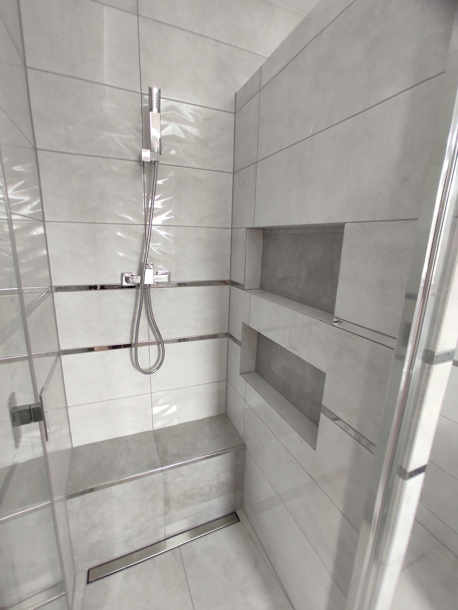 Nowoczesny prysznic z szarymi płytkami, chromowanymi listwami i wnękami w ścianie, odpływ liniowy w podłodze, widoczny fragment szklanej kabiny.