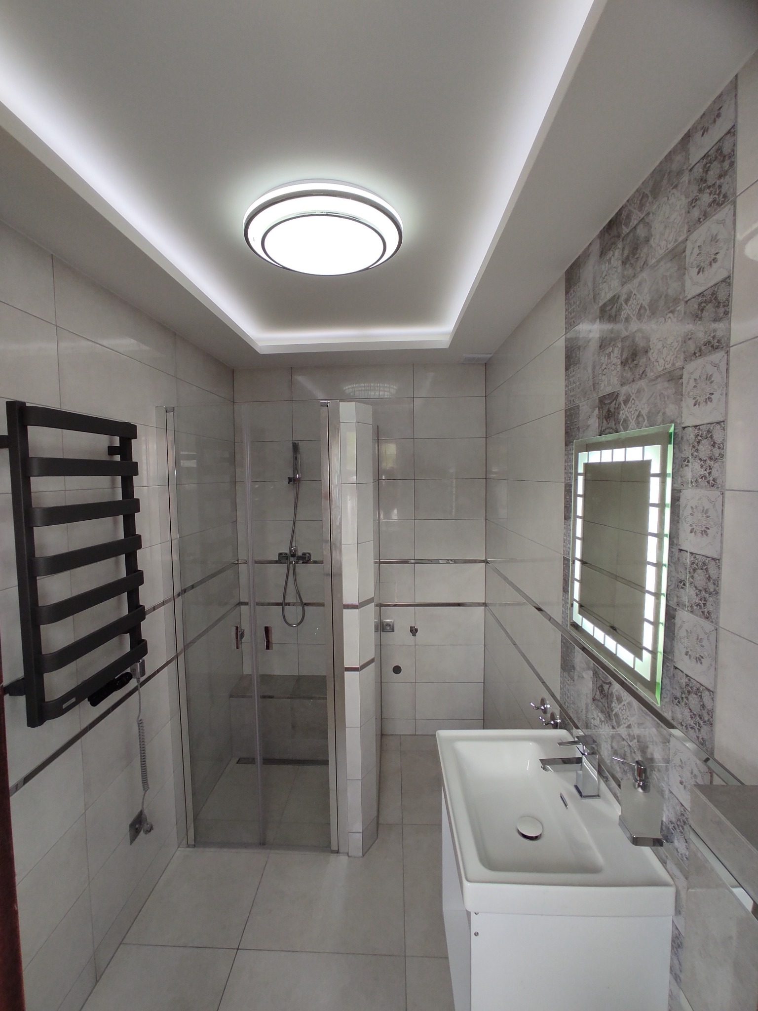 Nowoczesna łazienka z kabiną prysznicową, podświetlanym lustrem i grzejnikiem drabinkowym w kolorze czarnym.