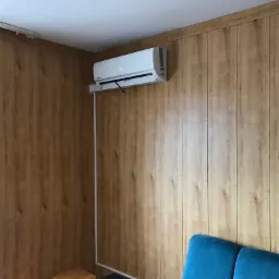 Biała klimatyzacja ścienna marki Kaisai zamontowana na ścianie wyłożonej panelami imitującymi drewno, widoczny niebieski fragment sofy.