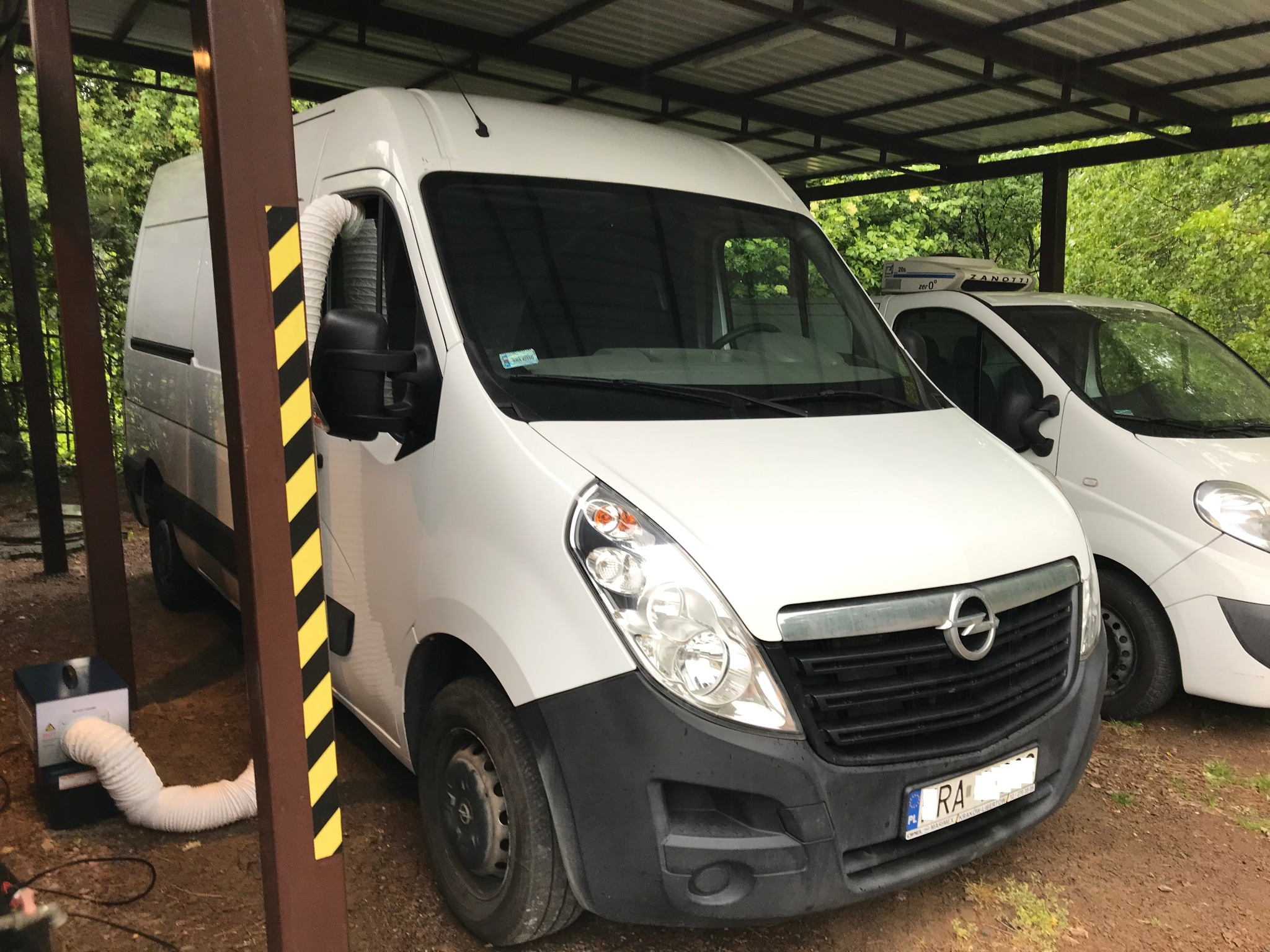 Biały van pod zadaszeniem z podłączonym urządzeniem z elastycznym przewodem w oknie, prawdopodobnie do dezynfekcji lub ozonowania.