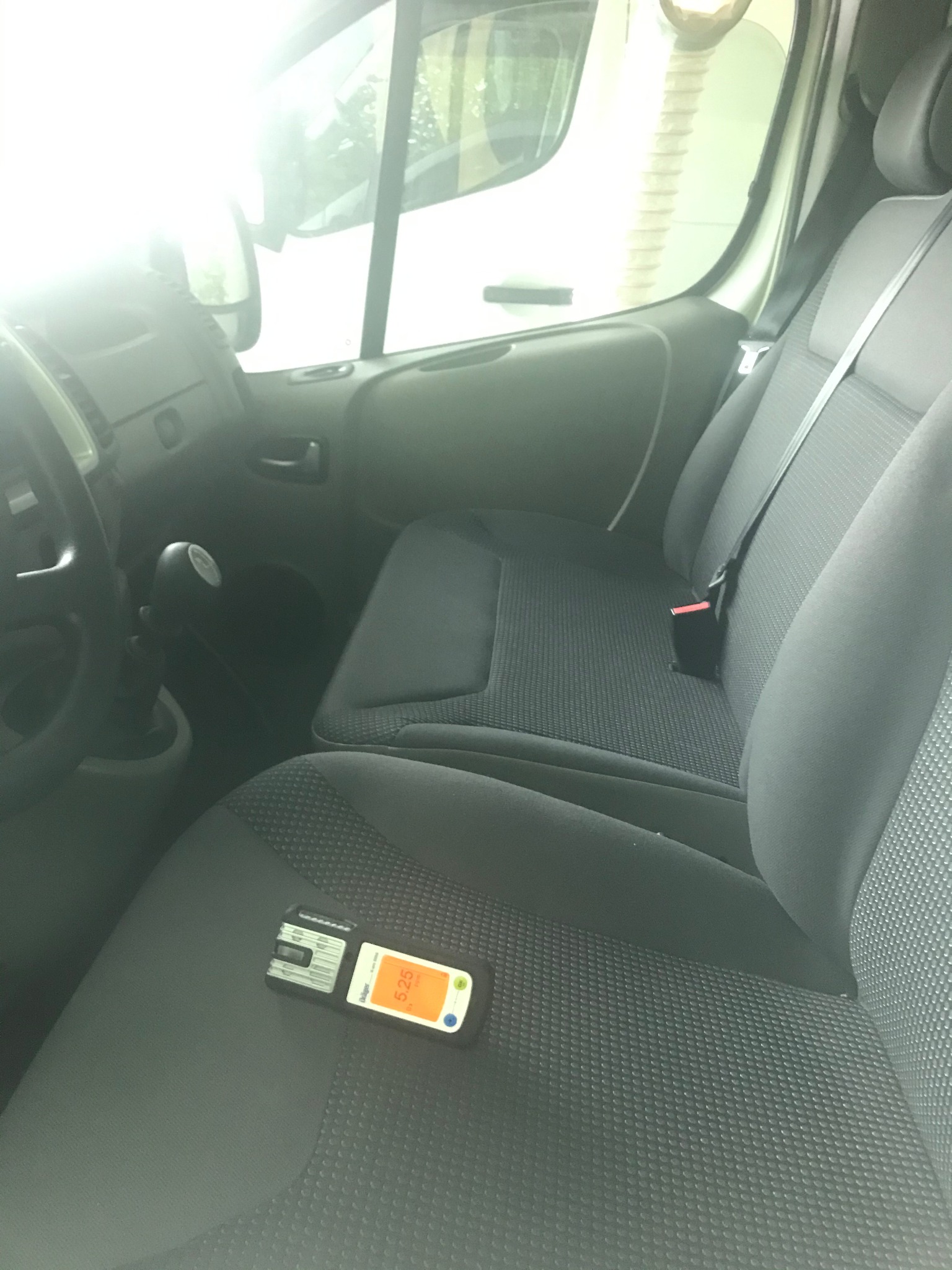 Miernik wielkości gazu na szarym siedzeniu samochodu dostawczego, widoczny kokpit i fragment białego samochodu przez szybę.