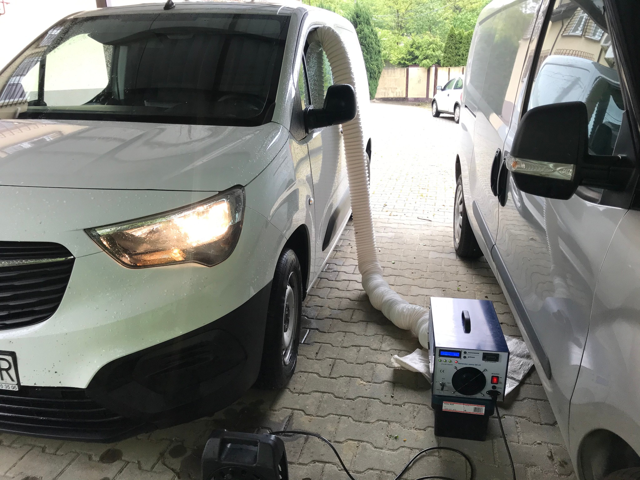 Biały samochód dostawczy z podłączonym elastycznym przewodem wentylacyjnym do generatora ozonu DG 32 stojącego na kostce brukowej, proces odkażania wnętrza pojazdu.