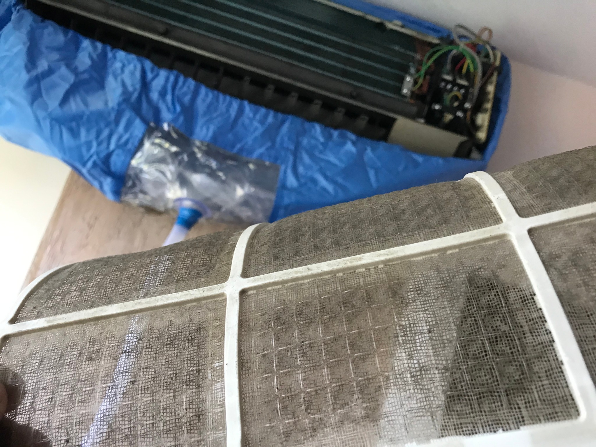 Brudny filtr powietrza klimatyzatora na pierwszym planie, w tle zabezpieczony folią klimatyzator podczas czyszczenia.