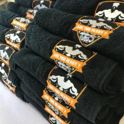 Stos czarnych ręczników z haftowanym logo siłacza i napisem 'Are you ready?' w pomarańczowej ramce, ułożonych na białym tle.