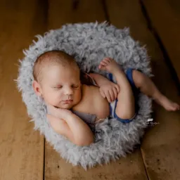 Śpiący noworodek w niebieskich spodenkach na puszystym, szarym kocyku w drewnianej misie, z delikatnym cieniem na twarzy.
