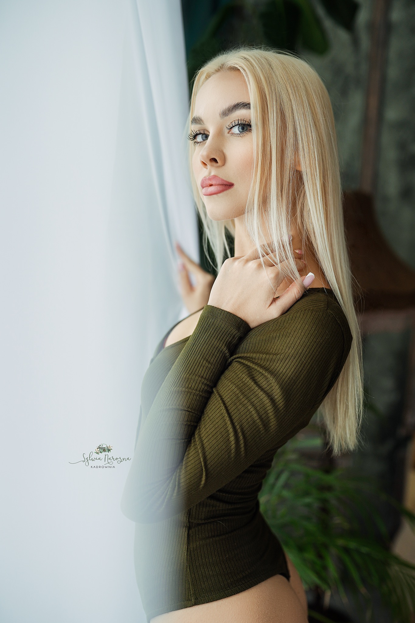Portret kobiety z blond włosami, w prążkowanym body w odcieniu khaki, stojącej przy oknie z białymi zasłonami, z delikatnym makijażem i wzrokiem skierowanym w bok, z widocznym logo fotografa...