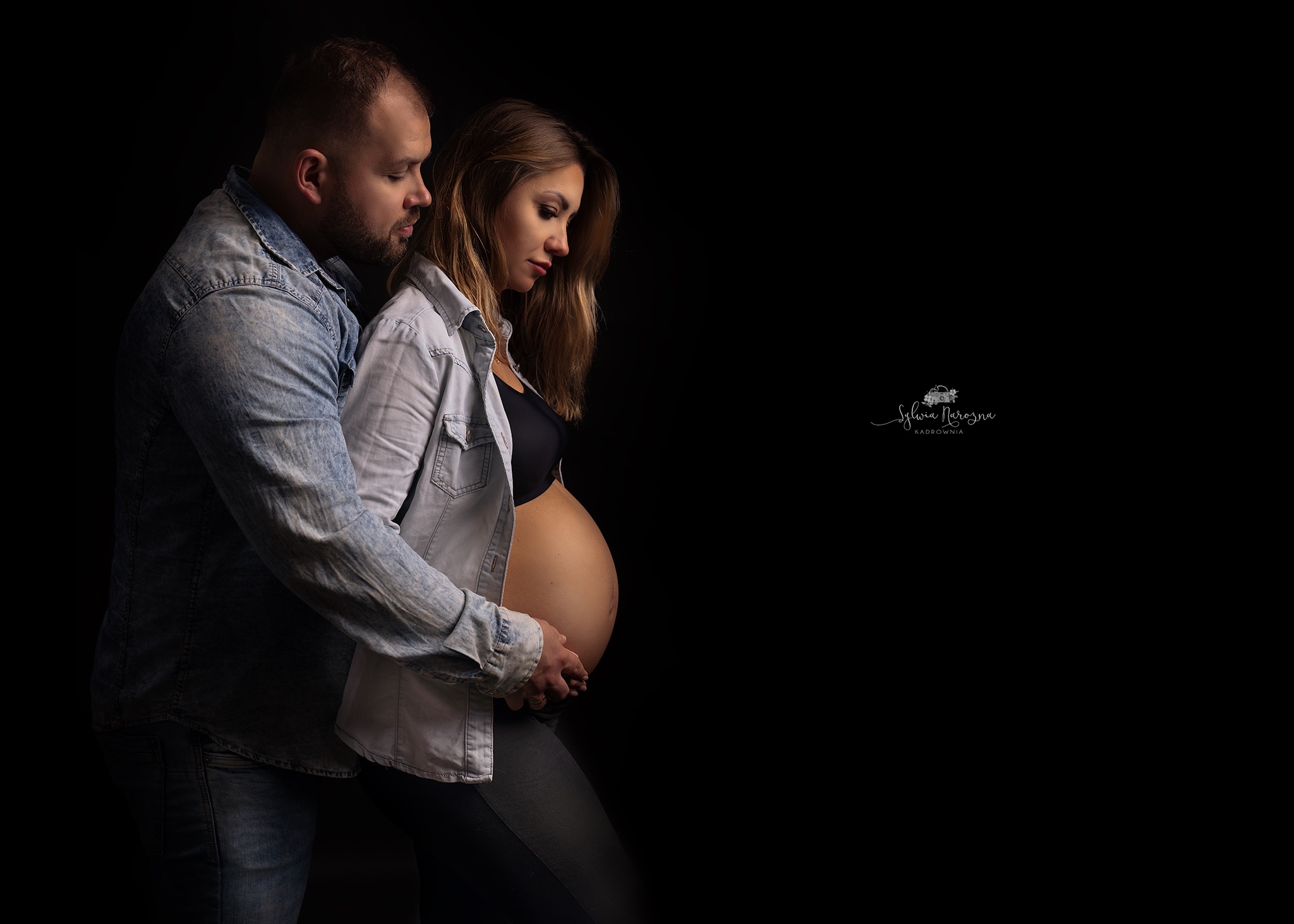 Przyszła mama z partnerem, oboje w dżinsowych koszulach, na czarnym tle, mężczyzna obejmuje ciążowy brzuch kobiety.