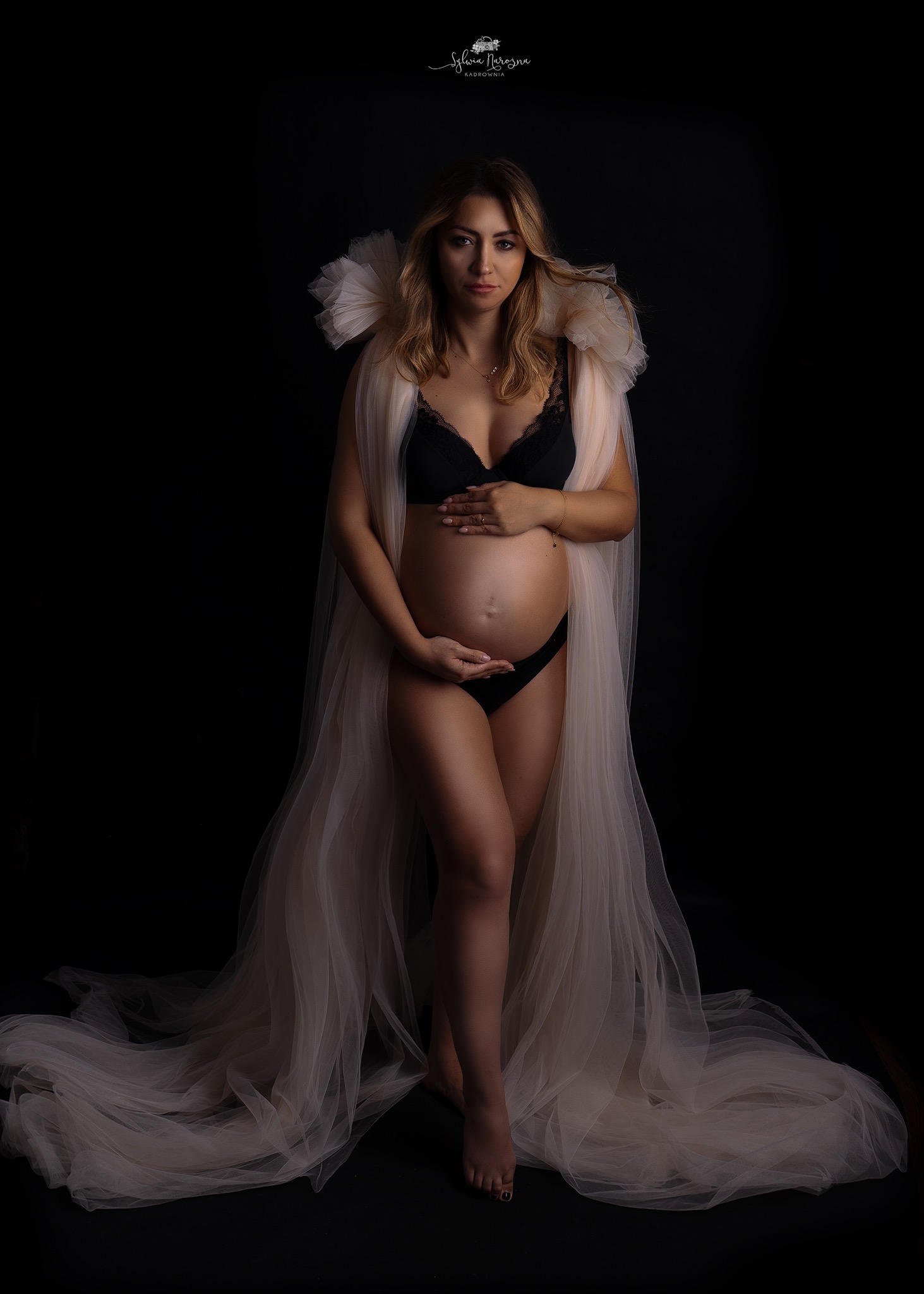 Kobieta w ciąży pozuje na czarnym tle, ubrana w czarną bieliznę i zwiewny, beżowy materiał z falbanami wokół szyi, trzyma ręce na brzuchu.