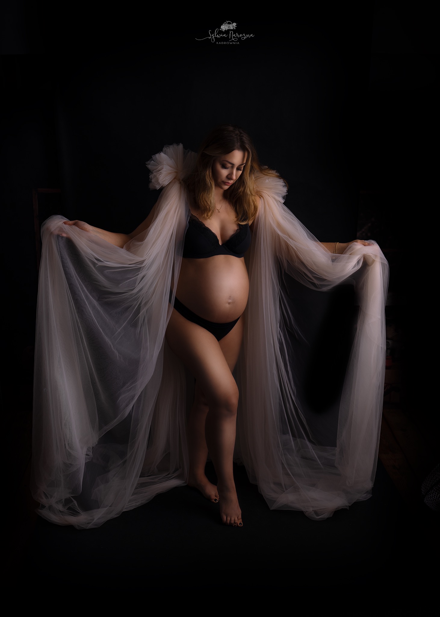 Kobieta w ciąży w czarnej bieliźnie, otulona przezroczystym, zwiewnym materiałem z bufkami na ramionach, pozuje na ciemnym tle.