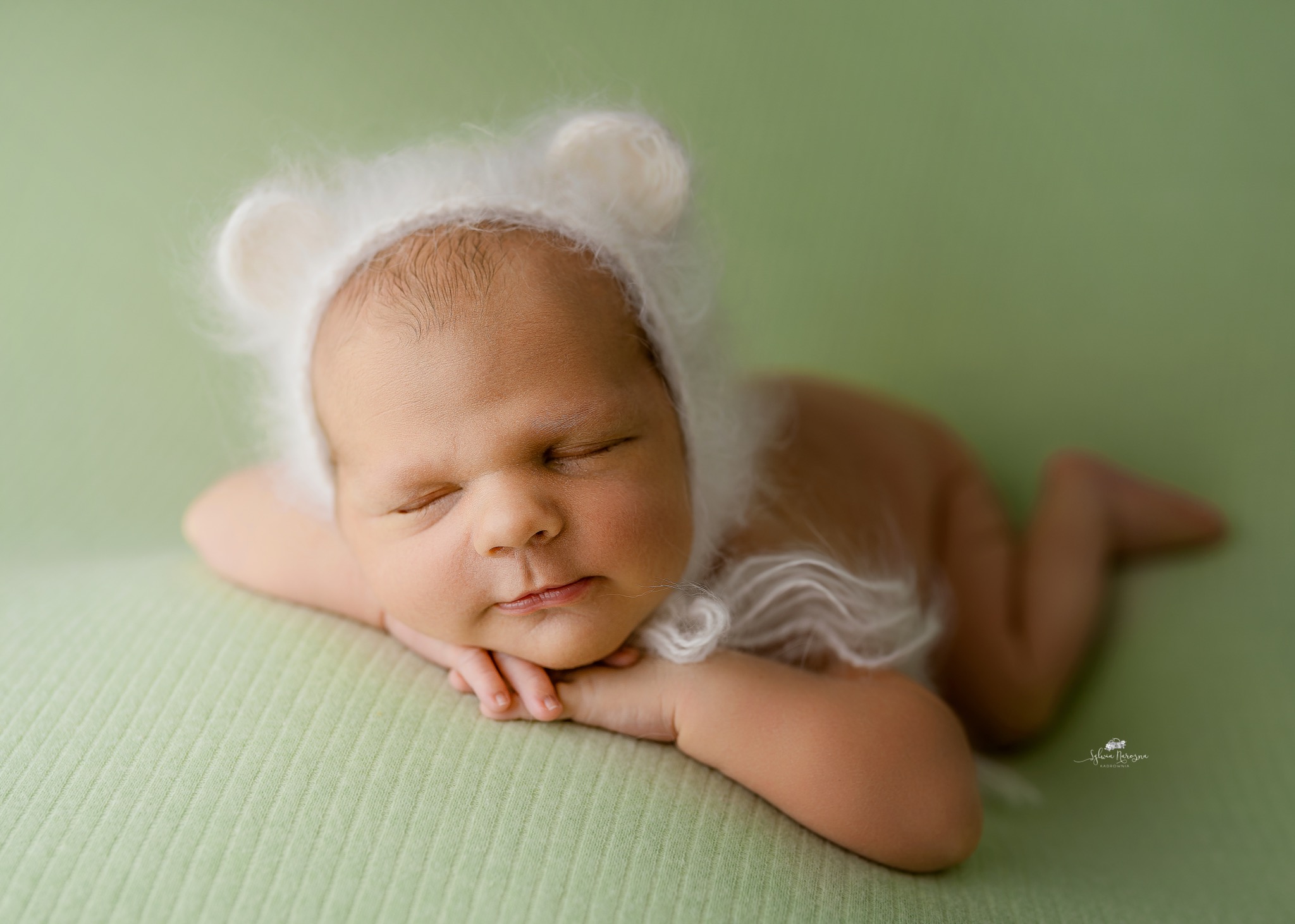Śpiący noworodek w puszystej czapce z uszkami, leżący na zielonym, prążkowanym kocyku, z rączkami pod policzkiem.