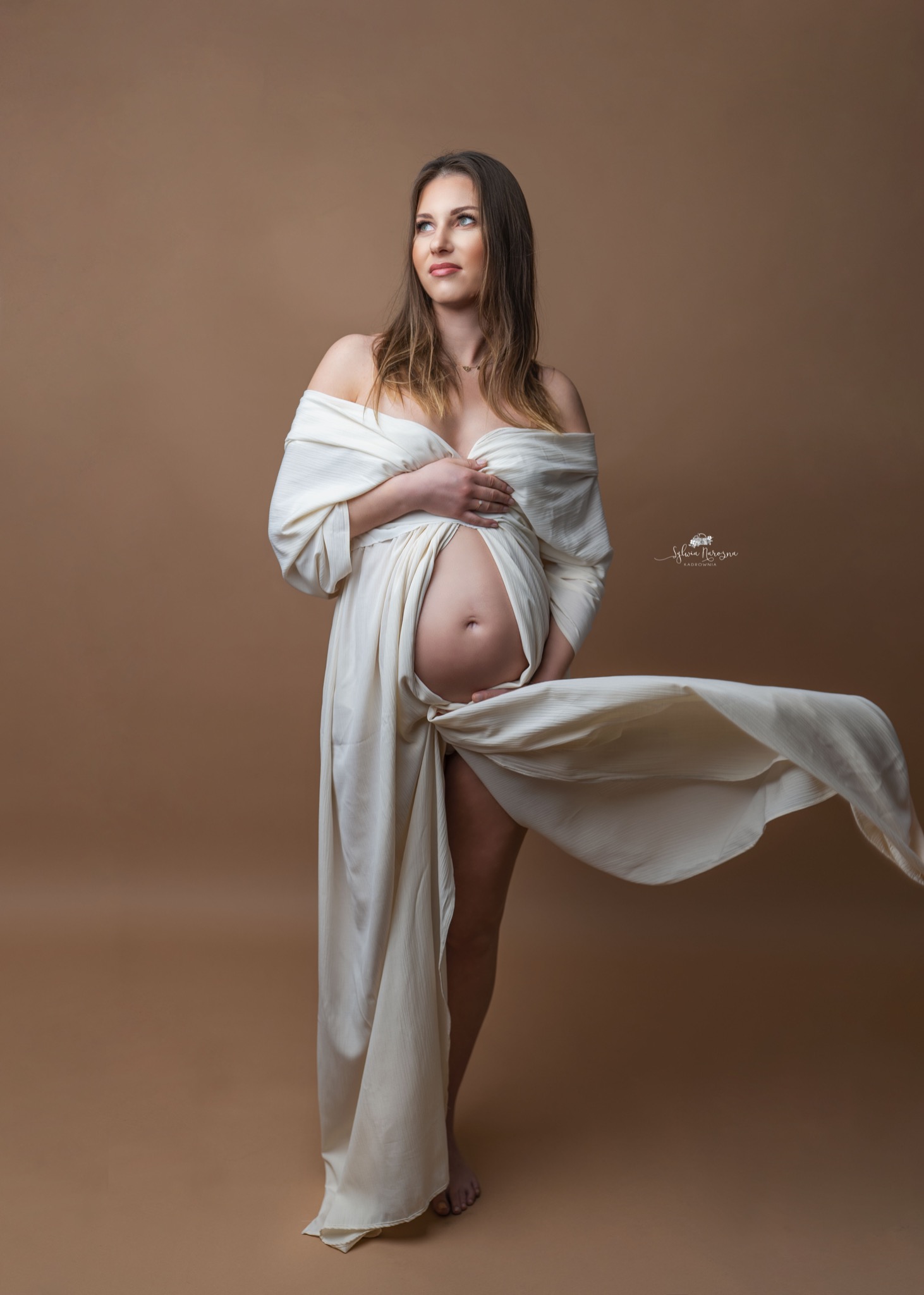 Kobieta w ciąży w białej, zwiewnej sukni pozuje na jednolitym, beżowym tle, spoglądając w górę w lewo; fragment sukni unosi się, tworząc dynamiczny efekt.