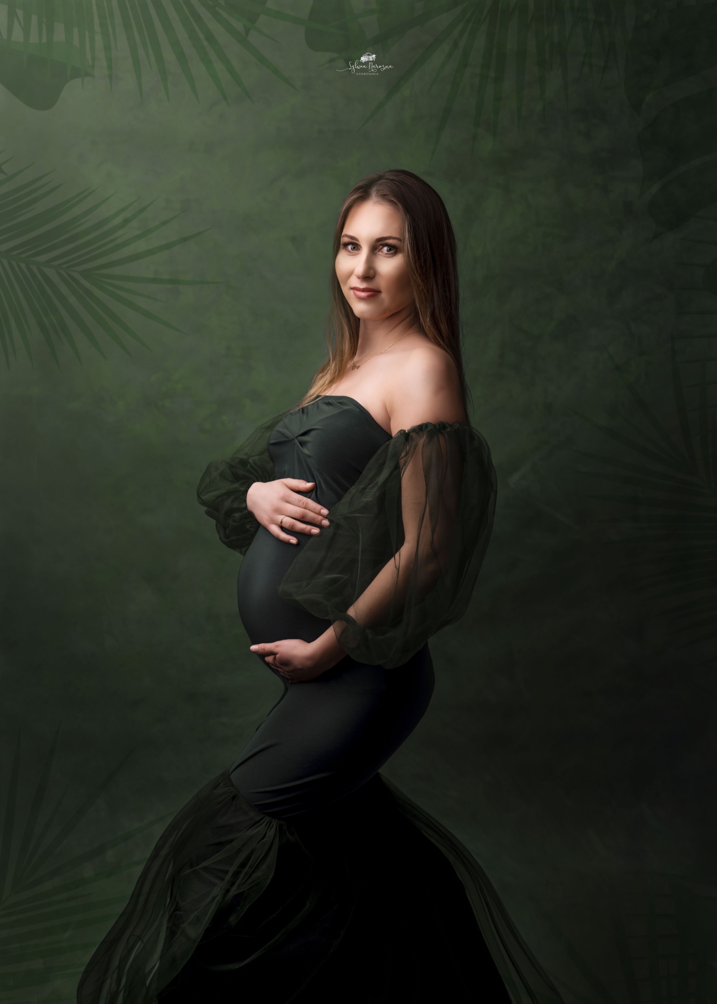 Portret kobiety w ciąży w długiej, eleganckiej sukni z bufiastymi rękawami na tle zielonego, teksturowanego tła z motywem liści palmowych.