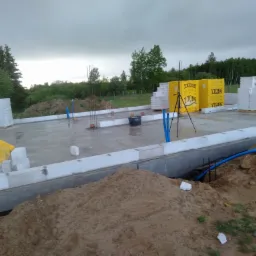 Fundament domu z betonu komórkowego Ytong na etapie budowy, widoczne zbrojenia, żółte paczki materiałów i hałdy ziemi w tle.