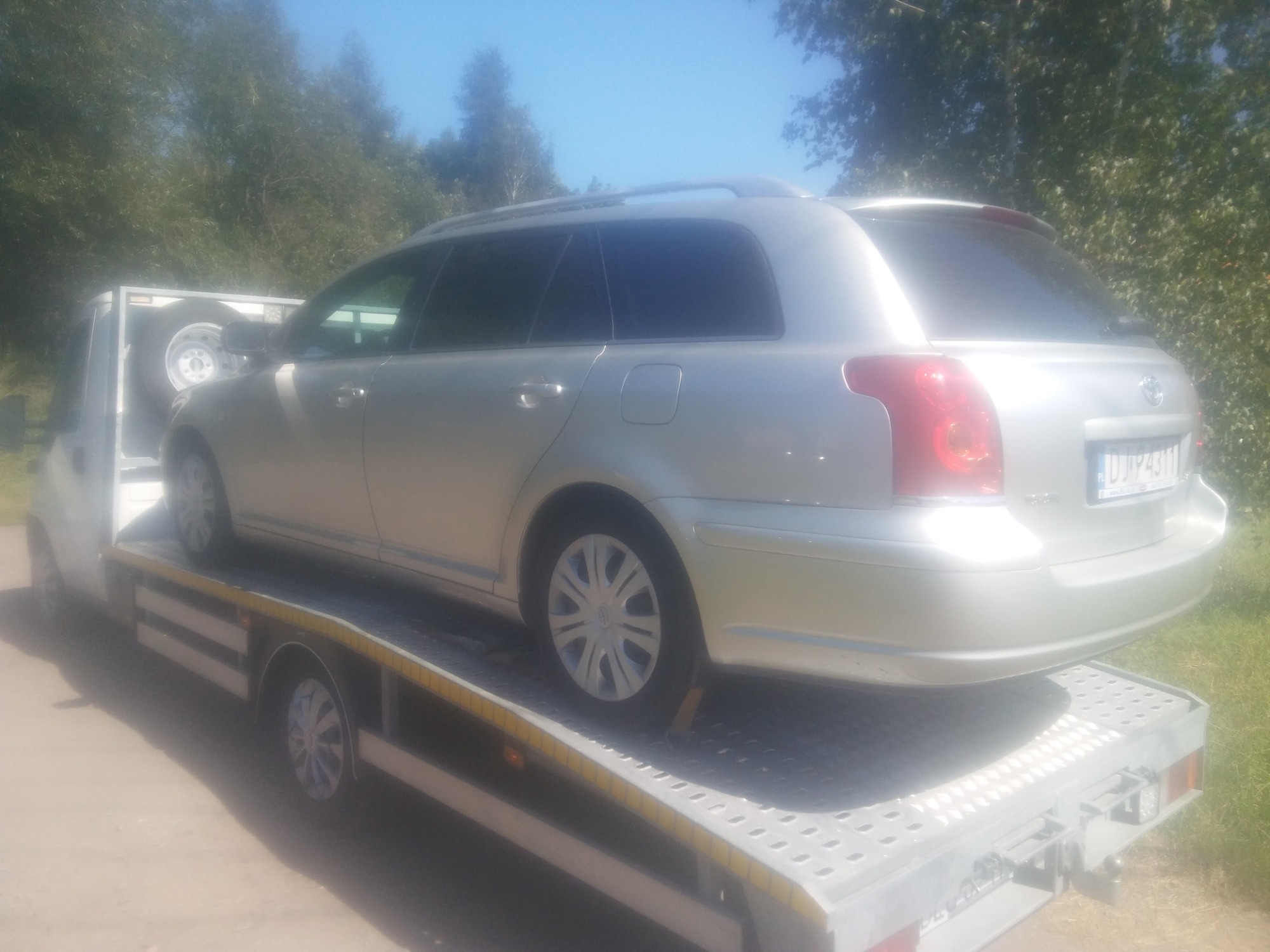 Srebrny samochód kombi marki Toyota na platformie lawety, widok z tyłu, dodatkowe koło zapasowe na lawecie, tablice rejestracyjne widoczne.