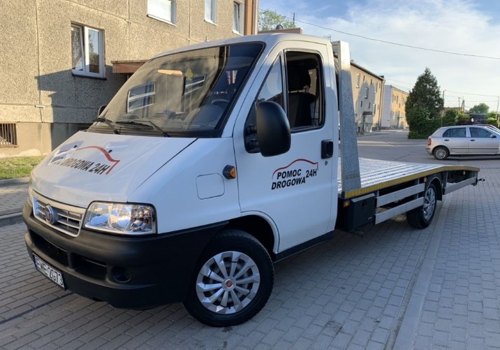 Biała laweta Fiat z napisem 'Pomoc Drogowa 24h' zaparkowana na chodniku przed budynkiem mieszkalnym.